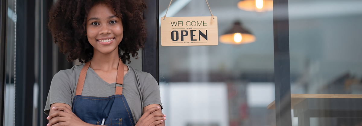 Smiling small business owner stands in front of store with open for business sign
